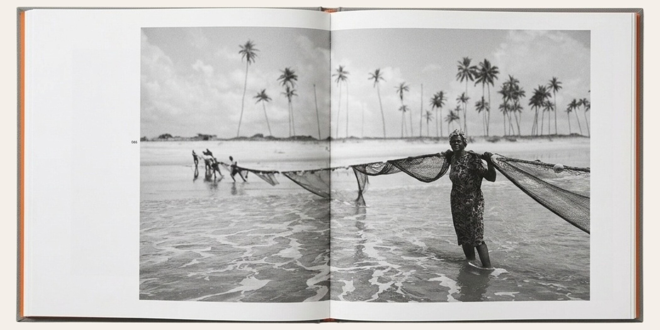 The Matriarch's Line opened flat to spread 086 — a wide black-and-white photograph of a woman in a floral dress standing knee-deep in the surf holding a fishing net, with a small crew hauling the same net behind her, tall palm trees along the horizon.