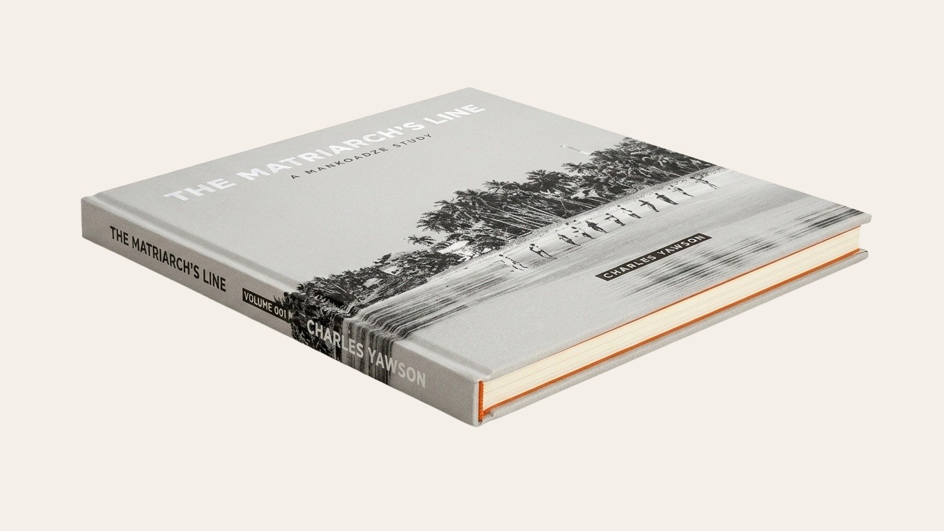The Standard Edition of The Matriarch's Line, paper-over-boards hardcover, closed, elevated 3/4 view. Wraparound black-and-white photograph — palm trees over a line of net-haulers on a wet Mankoadze beach — with foil-stamped title and Charles Yawson's name at the bottom. Orange endpapers visible along the fore-edge.